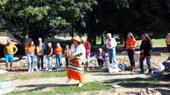 A person in traditional Indigenous regalia dances with a drum in front of a group of people during an outdoor cultural gathering.